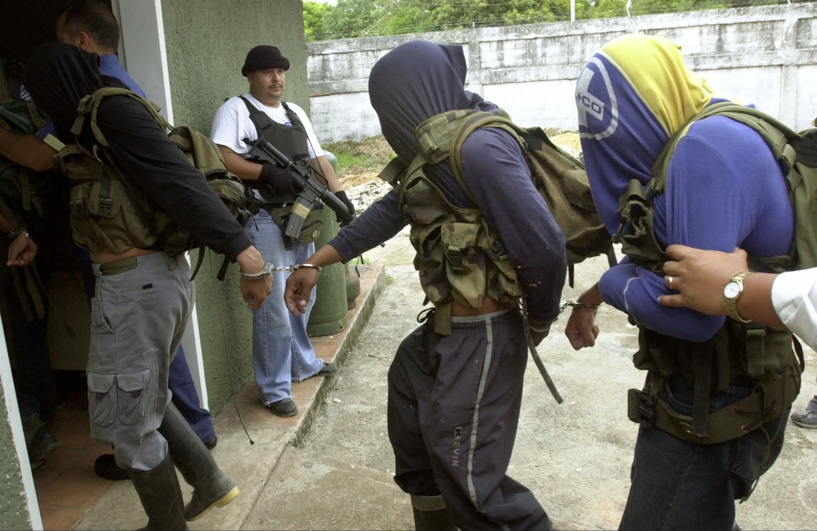 three men, handcuffed to each other and with their faces covered by their own T shirts, are led into a building. An armed man stands at the door