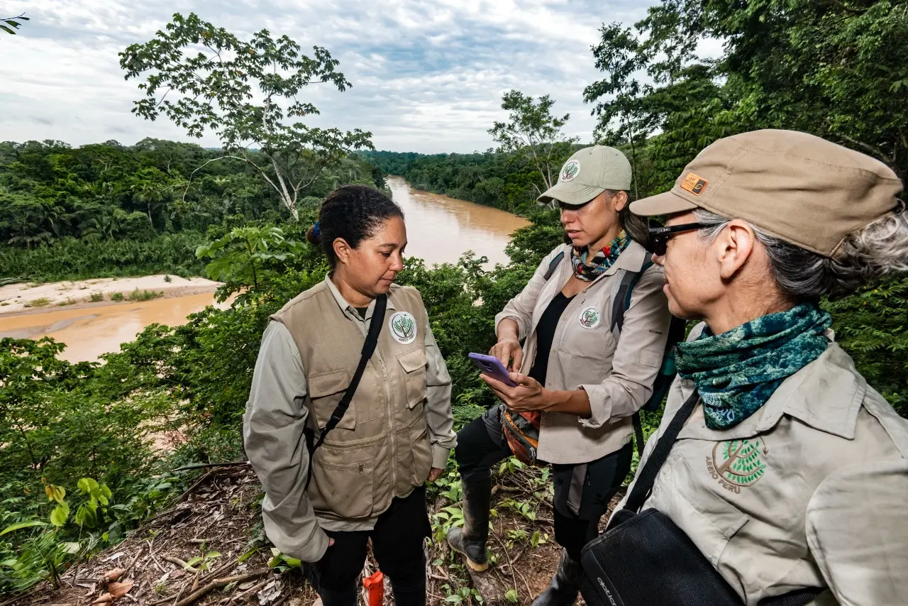 Three women in beige uniforms stand in the jungle by the river, conversing over a smartphone.
